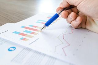 A close-up of a hand with a pen analyzing data on colorful bar and line charts on paper.