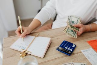 Adult holding cash and writing in planner while using a calculator at home.